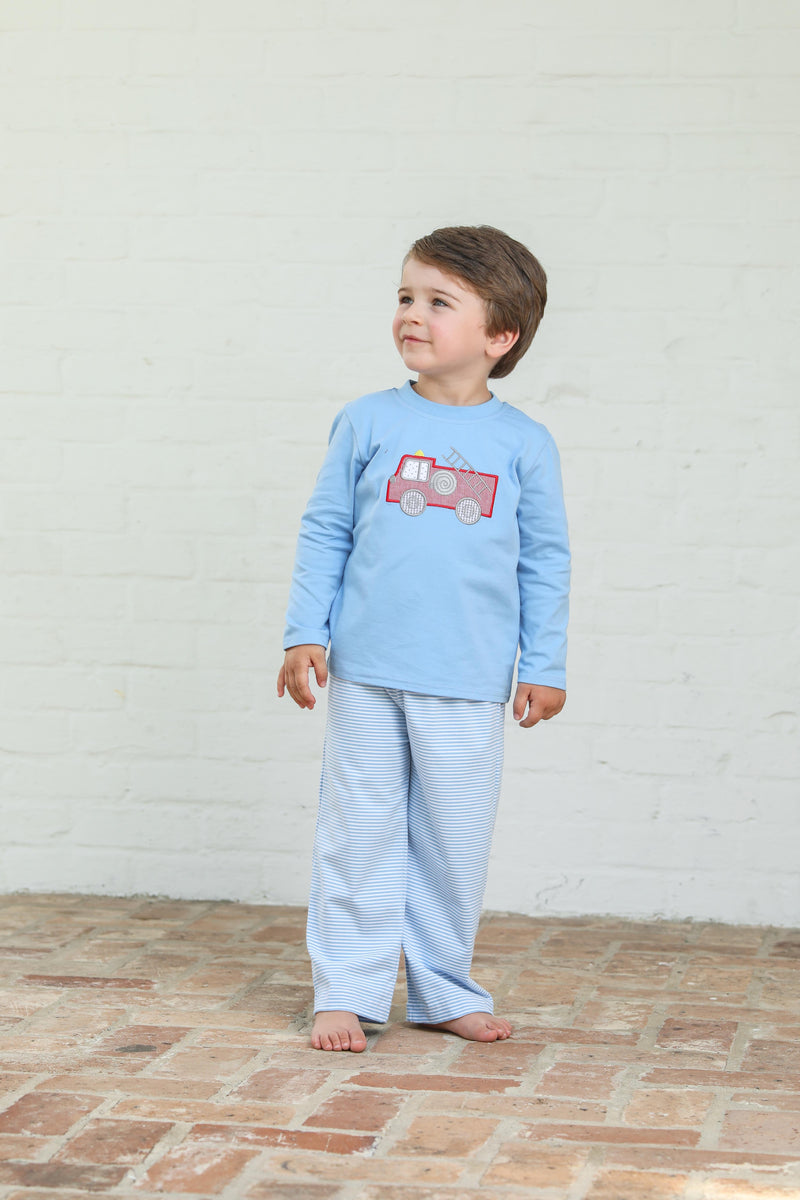 Child wearing a blue set with a red fire truck design against a white brick wall.