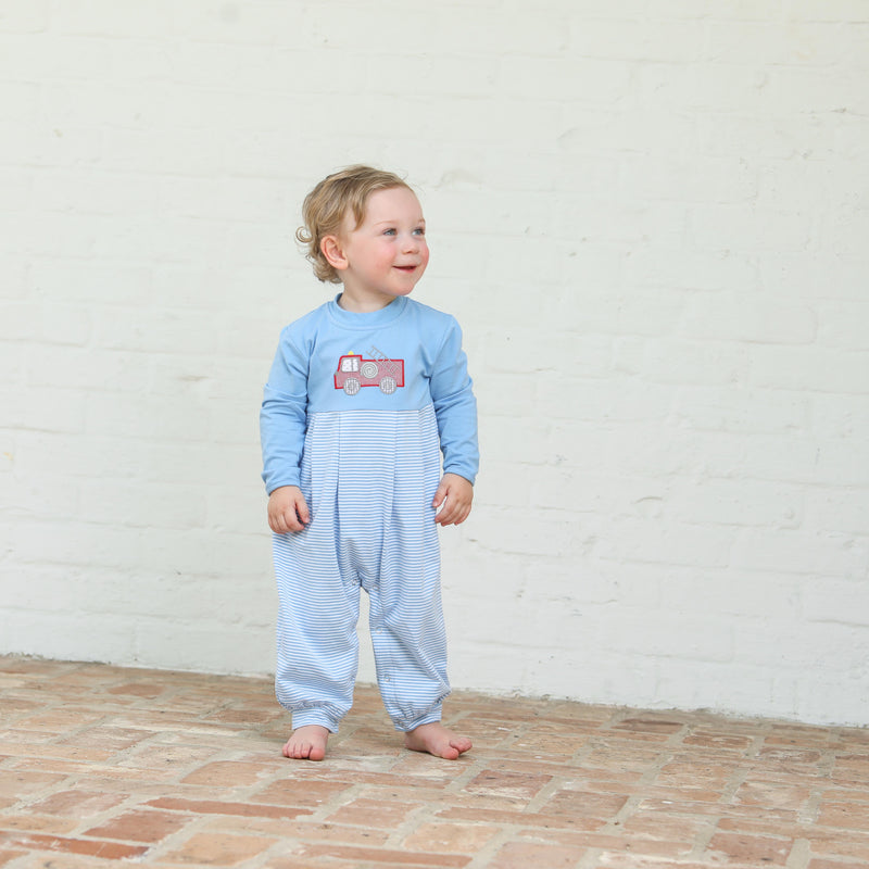 Child wearing a blue onesie jumpsuit with a red firetruck design against a white brick wall.